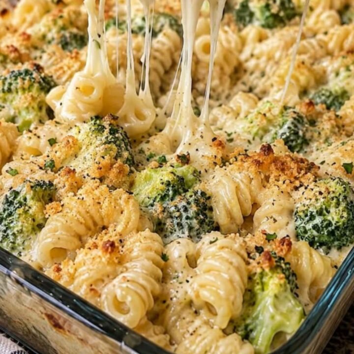 A close-up of a cheesy broccoli pasta bake in a glass dish with a long, stretchy cheese pull.