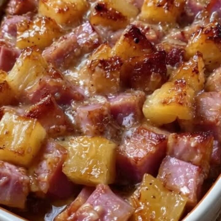 A white ceramic baking dish filled with caramelized, glistening ham and pineapple cubes coated in bubbling brown sugar glaze.