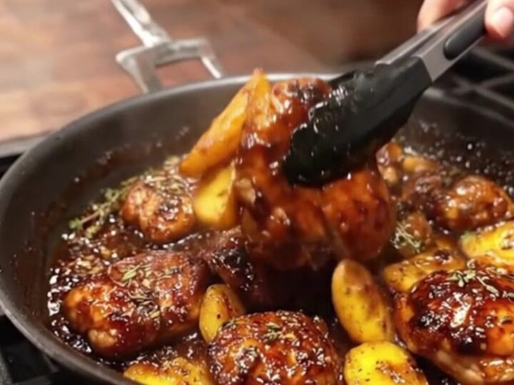 A close-up view of a cast-iron skillet filled with golden-brown chicken thighs and potato wedges coated in a glossy