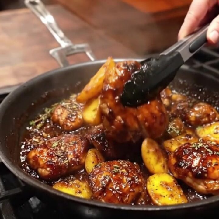 A close-up view of a cast-iron skillet filled with golden-brown chicken thighs and potato wedges coated in a glossy