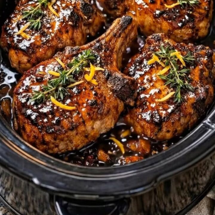 Close-up of a tender, glazed bone-in pork chop sitting in a slow cooker with a rich, aromatic orange herb sauce.