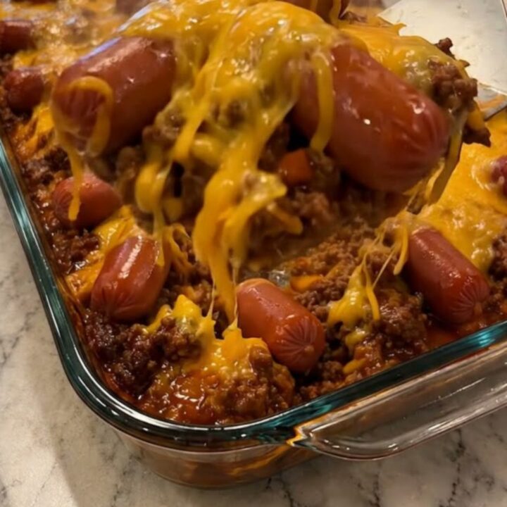 A metal spatula lifting a steaming scoop of chili cheese dog casserole from a glass dish.