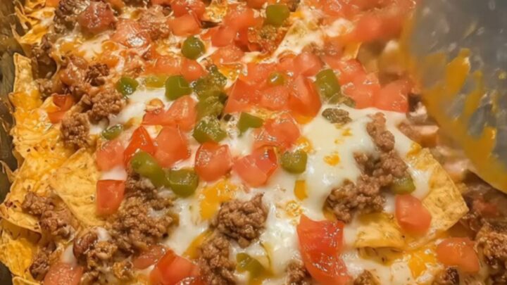 A close-up of a loaded beef nacho bake in a glass dish with a generous, gooey cheese pull held by a metal spatula.