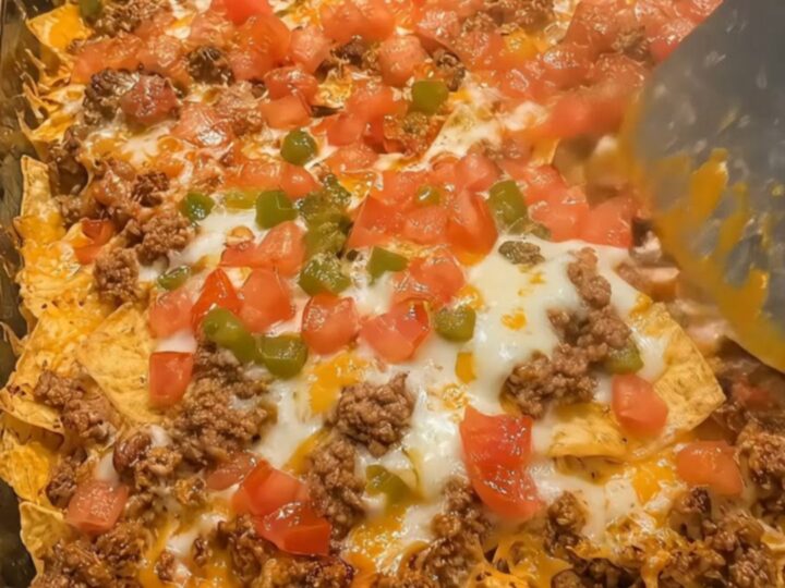 A close-up of a loaded beef nacho bake in a glass dish with a generous, gooey cheese pull held by a metal spatula.