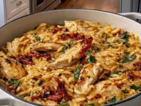 A close-up of a creamy chicken orzo dish in a white enameled braiser, featuring visible sun-dried tomatoes and spinach.