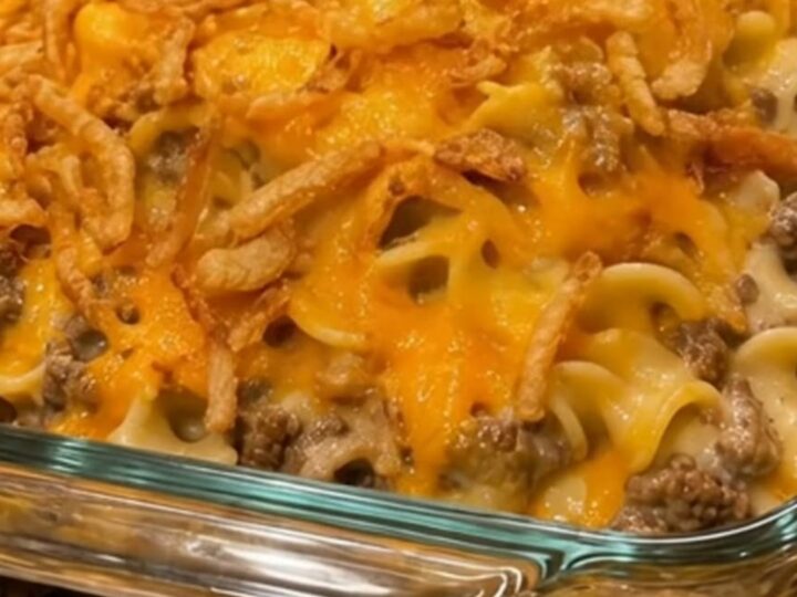A metal spoon scoops a steaming portion of cheesy beef and noodle casserole from a glass baking dish.