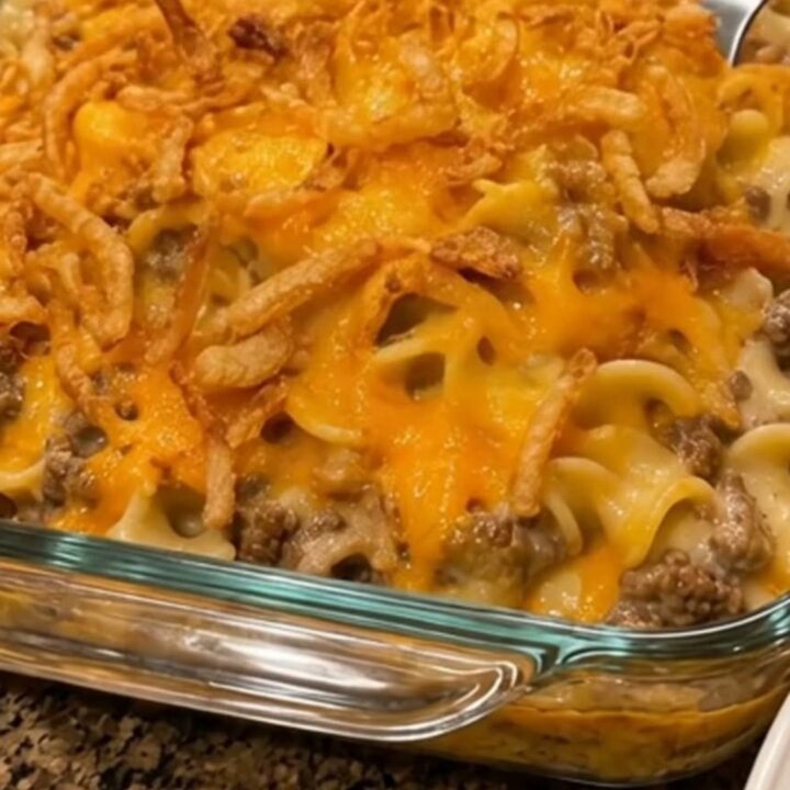 A metal spoon scoops a steaming portion of cheesy beef and noodle casserole from a glass baking dish.
