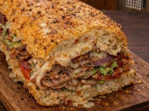 A close-up of a loaded Italian cheese melt sandwich on a wooden cutting board with a visible cheese pull.