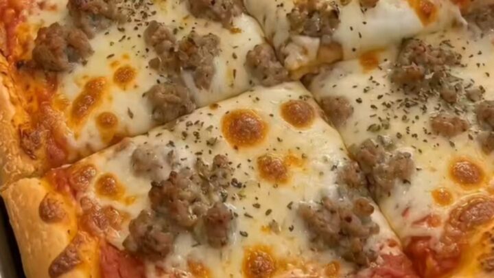 A golden-brown, rectangular sheet pan pizza topped with melted cheese and crumbled sausage.