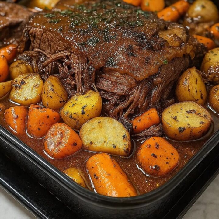 A close-up of a tender beef chuck roast surrounded by golden potatoes and carrots in a rich gravy inside a roasting dish.