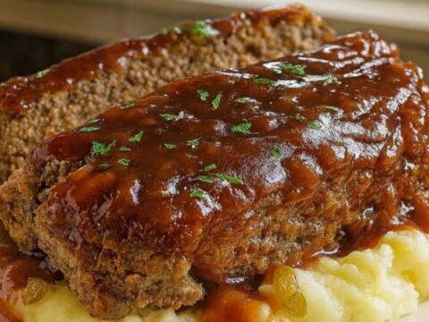 A thick glazed slice of tender Mississippi meatloaf resting over fluffy mashed potatoes on a white plate.