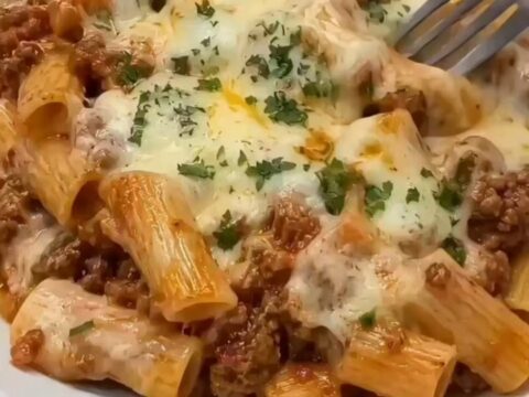 A steaming, freshly baked beef rigatoni pasta in a ceramic dish, topped with golden melted cheese and fresh parsley.