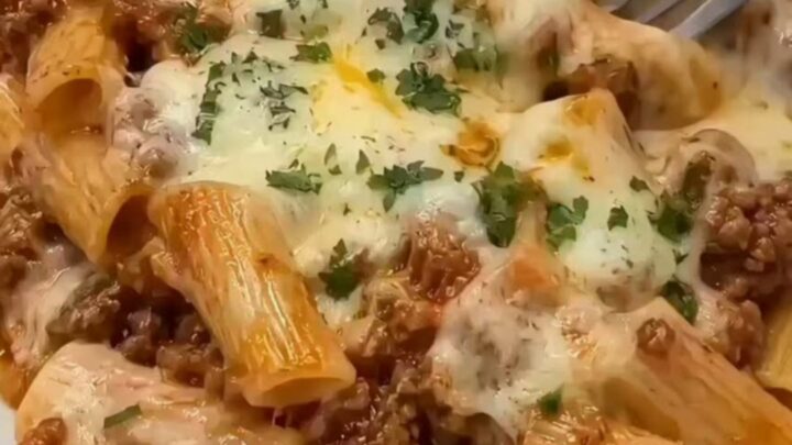 A steaming, freshly baked beef rigatoni pasta in a ceramic dish, topped with golden melted cheese and fresh parsley.