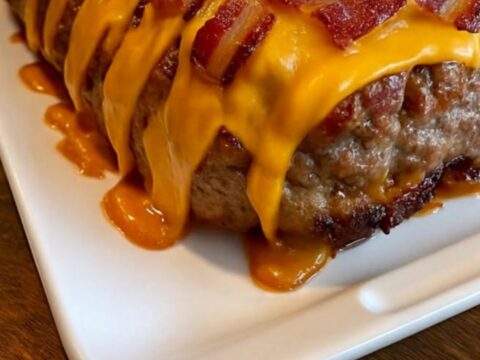 A thick, steaming slice of bacon cheeseburger meatloaf topped with melted cheddar and crispy bacon on a wooden counter.