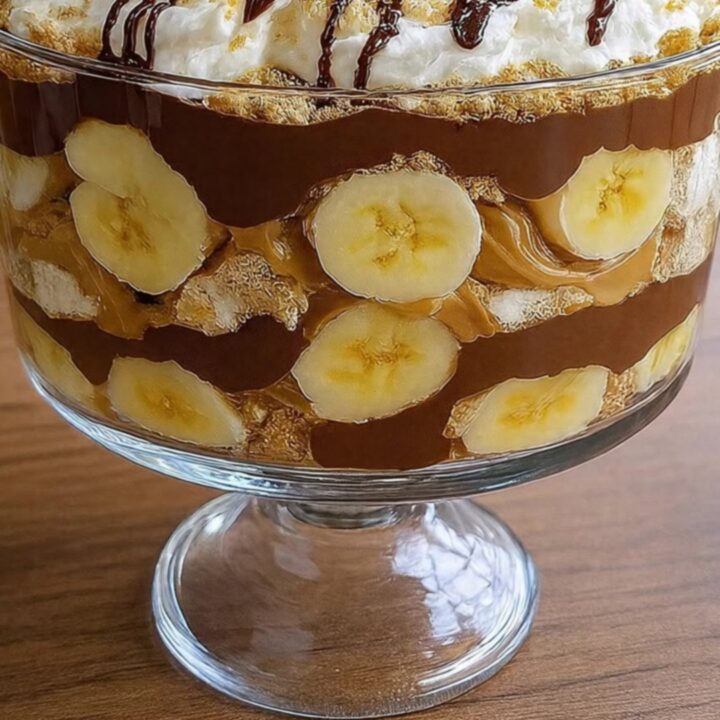 A glass pedestal bowl filled with layers of chocolate pudding, peanut butter cream, banana slices, and graham crumbs.