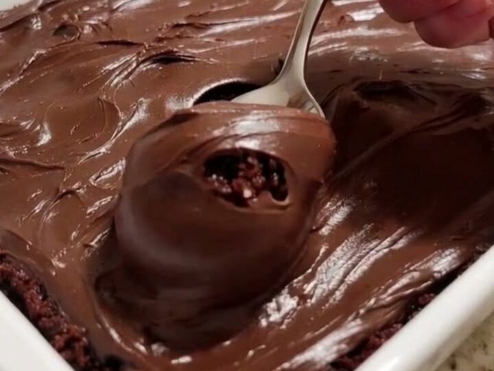 A slice of moist, dark chocolate cake topped with thick chocolate fudge frosting on a white plate.