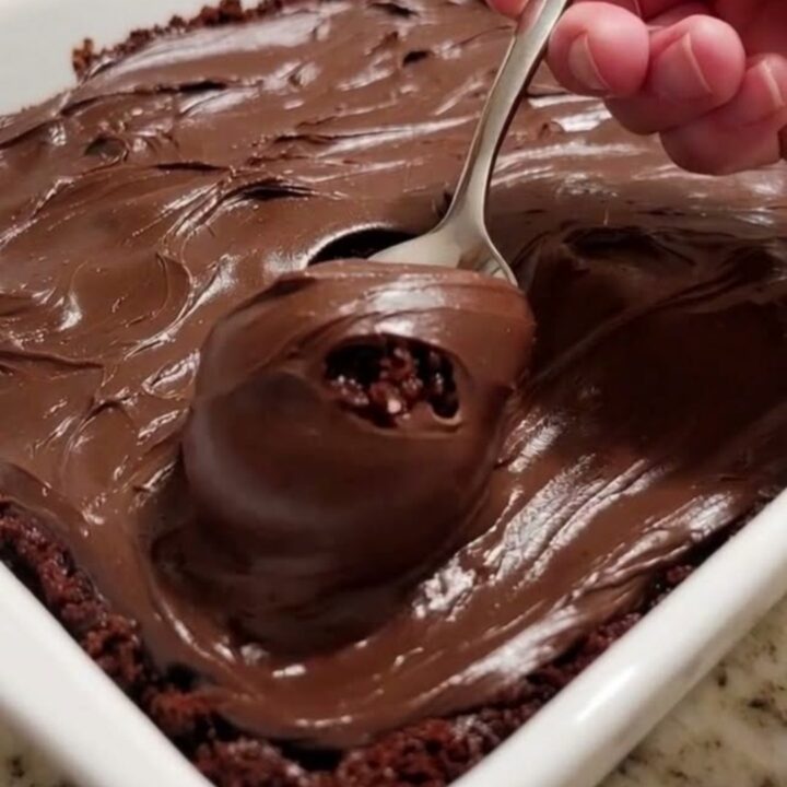 A slice of moist, dark chocolate cake topped with thick chocolate fudge frosting on a white plate.