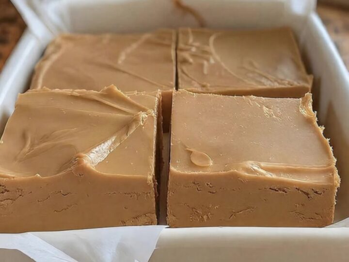 A close-up of a square of peanut butter fudge being lifted from a ceramic dish with a small metal spatula.