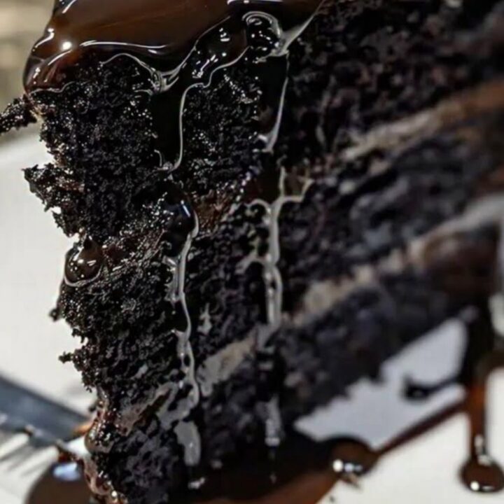 A slice of black velvet cake on a white plate with a glossy chocolate ganache dripping down the side.