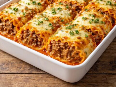 A close-up of a cheesy short rib lasagna roll being lifted from a white baking dish with a spatula.