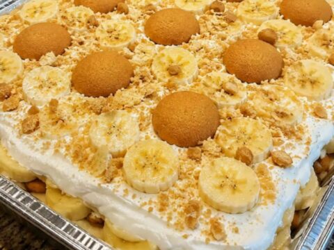 A large foil pan filled with layers of banana pudding, sliced bananas, and vanilla wafers topped with whipped cream.