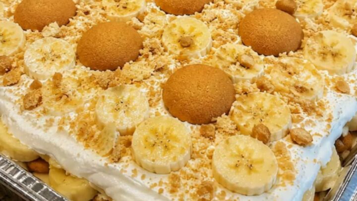 A large foil pan filled with layers of banana pudding, sliced bananas, and vanilla wafers topped with whipped cream.