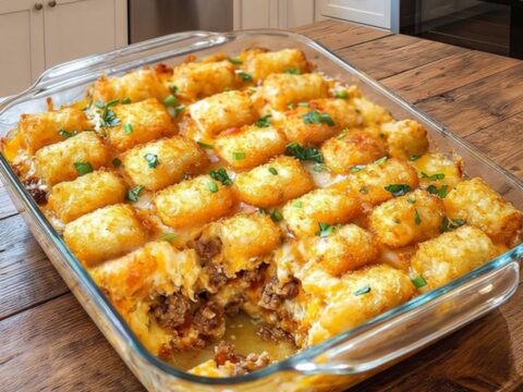 A glass baking dish filled with a golden, cheesy tater tot beef casserole sitting on a wooden kitchen counter.