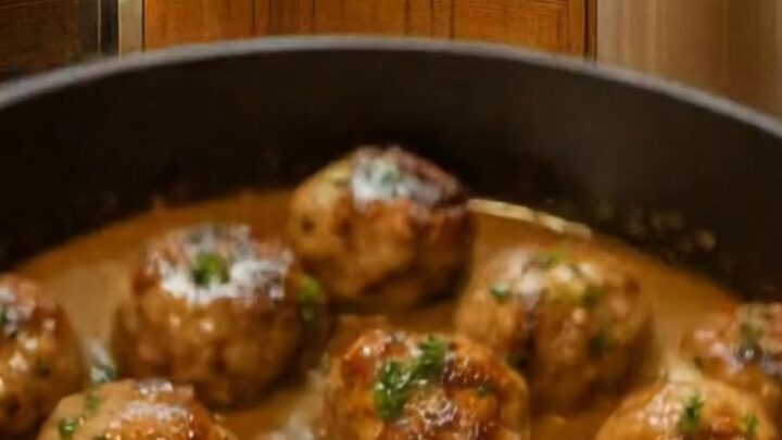 A close-up of glossy meatballs covered in a creamy sauce, garnished with fresh green parsley in a black skillet.