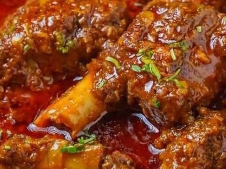 A close-up of tender, braised lamb shanks covered in a rich, spicy red gravy with fresh cilantro.