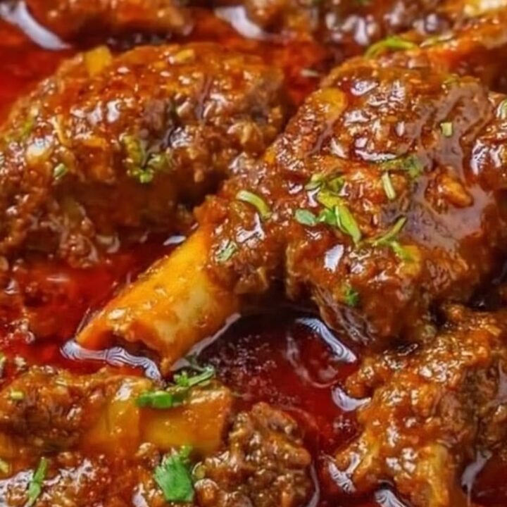 A close-up of tender, braised lamb shanks covered in a rich, spicy red gravy with fresh cilantro.