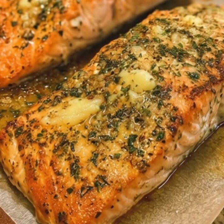 A perfectly baked salmon fillet with a golden, glistening garlic herb butter crust on a white plate.