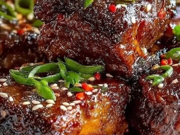 A close-up of glistening, dark glazed short ribs on a white serving platter, garnished with sesame seeds and green onions.