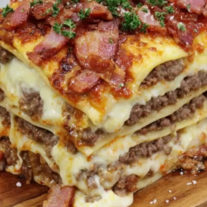 A tall, multi-layered slice of cheeseburger lasagna garnished with crispy bacon and chopped parsley.