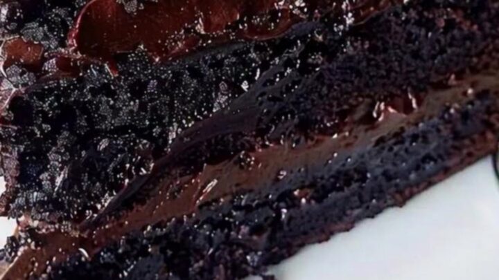 A slice of decadent dark chocolate fudge cake with glossy frosting on a white plate.