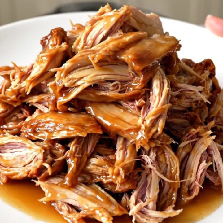 A close-up of tender, glossy apple cider pulled chicken served on a white plate.