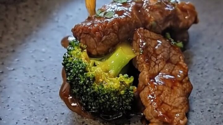 A close-up view of tender flank steak and bright green broccoli florets in a rich brown sauce on a grey plate.