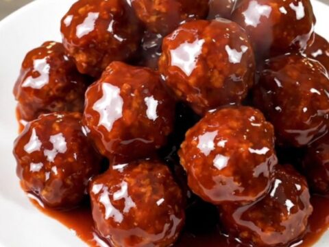 A close-up view of steaming, glossy meatballs covered in a thick, dark red glaze in a slow cooker.