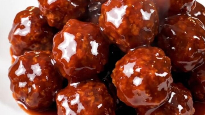 A close-up view of steaming, glossy meatballs covered in a thick, dark red glaze in a slow cooker.