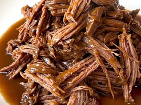 A close-up view of a tender shredded pot roast coated in a rich, glossy brown gravy on a white plate.