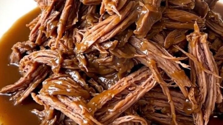 A close-up view of a tender shredded pot roast coated in a rich, glossy brown gravy on a white plate.