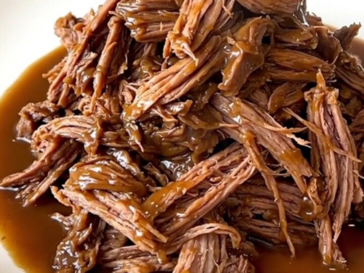 A close-up view of a tender shredded pot roast coated in a rich, glossy brown gravy on a white plate.