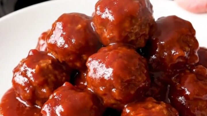 A top-down view of a black slow cooker filled with meatballs covered in a glossy cranberry chili sauce.