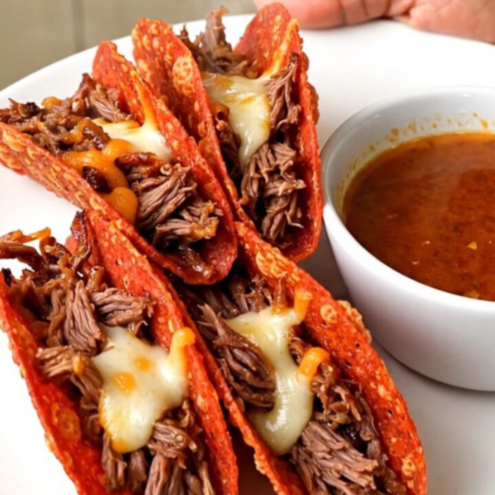A close-up of crispy, golden-brown beef birria tacos served on a white plate with a side of savory dipping broth.