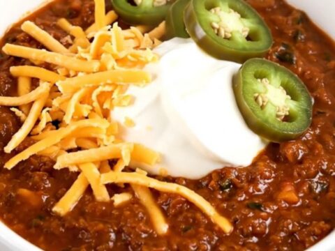 A steaming bowl of beef chili topped with cheddar cheese, sour cream, and jalapeño slices.