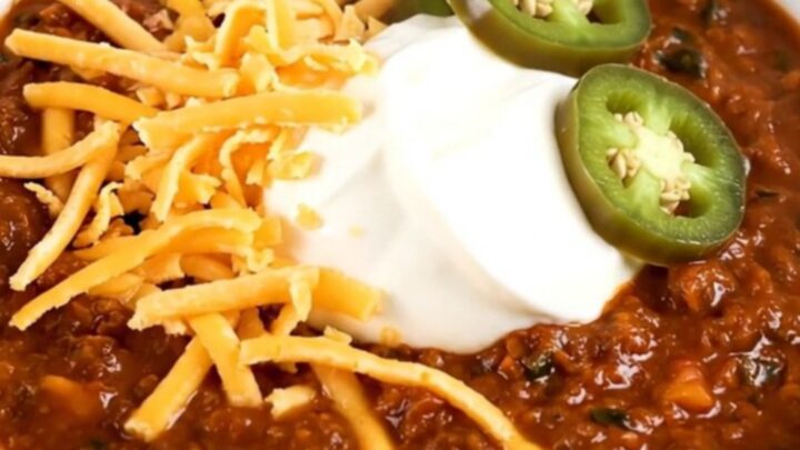 A steaming bowl of beef chili topped with cheddar cheese, sour cream, and jalapeño slices.