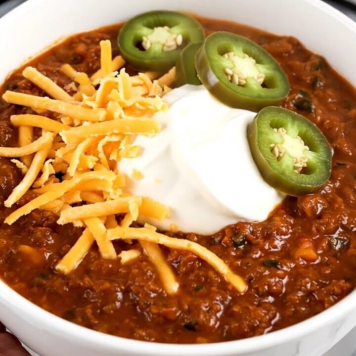 A steaming bowl of beef chili topped with cheddar cheese, sour cream, and jalapeño slices.