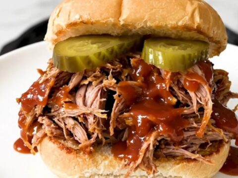 A close-up of a tender pulled pork sandwich on a soft bun with pickles.