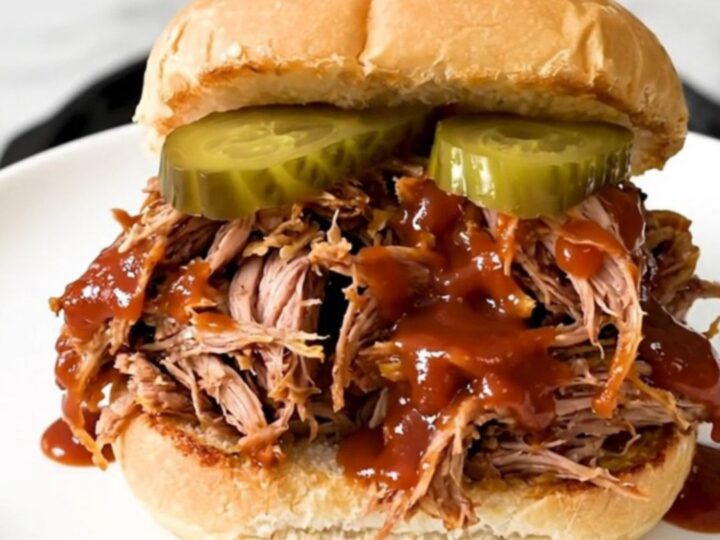 A close-up of a tender pulled pork sandwich on a soft bun with pickles.