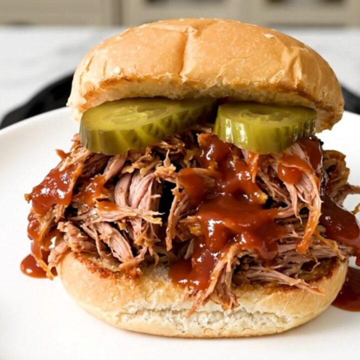 A close-up of a tender pulled pork sandwich on a soft bun with pickles.