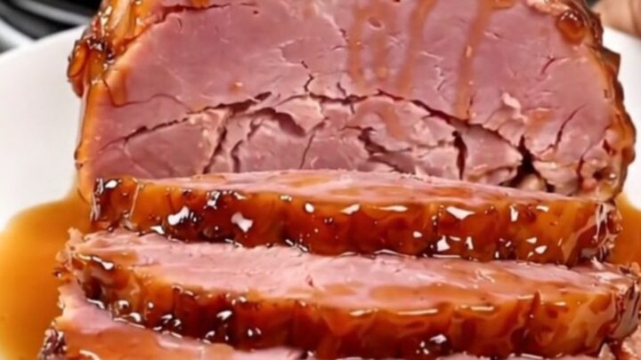 A succulent, glazed boneless ham sitting in a black slow cooker on a white speckled countertop.